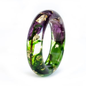 K&ouml;nnte beinhalten: Ein klarer Harz-Armreif mit eingebetteten lila Blumen, gr&uuml;nem Laub und Goldblatt-Akzenten. Das Armband hat eine glatte, abgerundete Form und ist vor einem wei&szlig;en Hintergrund platziert. Das florale Design ist im gesamten Armband sichtbar.