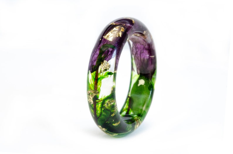 K&ouml;nnte beinhalten: Ein klarer Harz-Armreif mit eingebetteten lila Blumen, gr&uuml;nem Laub und Goldblatt-Akzenten. Das Armband hat eine glatte, abgerundete Form und ist vor einem wei&szlig;en Hintergrund platziert. Das florale Design ist im gesamten Armband sichtbar.