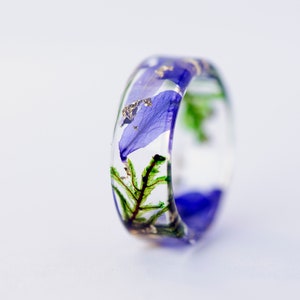 May include: A clear resin ring with blue flower petals, green foliage, and gold flakes embedded within. The ring has a rounded shape and is set against a white background.