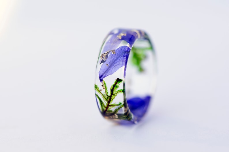 May include: A clear resin ring with blue flower petals, green foliage, and gold flakes embedded within. The ring has a rounded shape and is set against a white background.