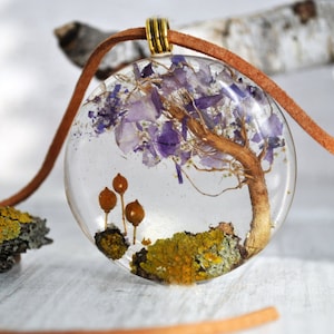 May include: A round, clear resin pendant with a brown leather cord. Inside the pendant is a small, dried tree with purple flowers and green moss.