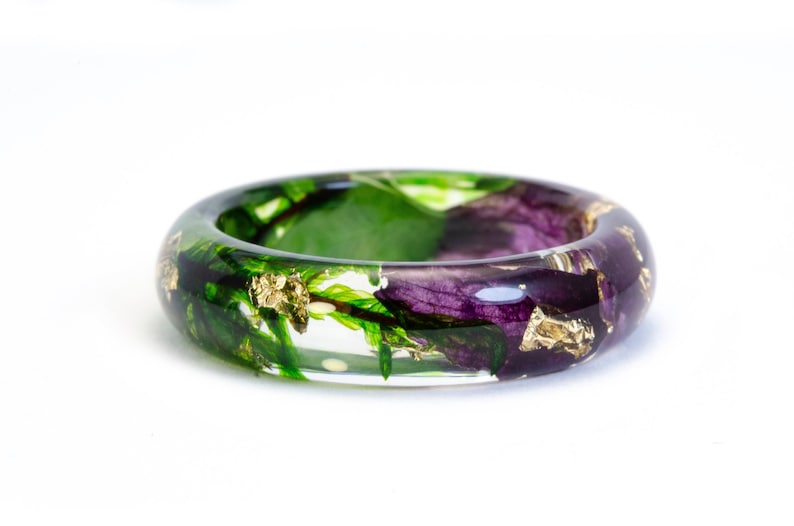 K&ouml;nnte beinhalten: Ein klarer Harzring mit eingebetteten gr&uuml;nen Bl&auml;ttern, lila Bl&uuml;tenbl&auml;ttern und Goldblattflocken. Der Ring ist ein dickes Band, das die eingeschlossenen Naturelemente zeigt. Das Design ist vor einem wei&szlig;en Hintergrund.