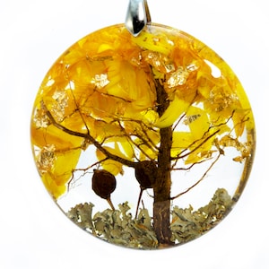 May include: A round pendant with a clear resin background and a small tree with yellow leaves and brown branches. The tree is surrounded by small, gray, lichen-like pieces.
