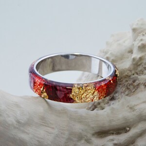 Puede incluir: Un anillo de banda de plata con una incrustaci&oacute;n de resina roja y dorada. La resina tiene un efecto marmoleado con motas de oro.
