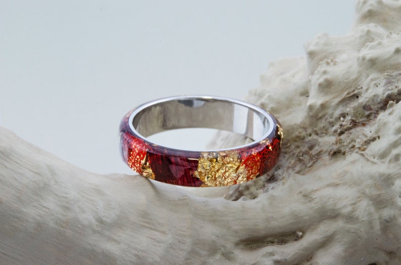 Puede incluir: Un anillo de banda de plata con una incrustaci&oacute;n de resina roja y dorada. La resina tiene un efecto marmoleado con motas de oro.