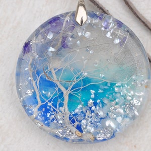May include: A round, clear resin pendant with a blue and white tree design. The pendant is filled with silver glitter and white and blue iridescent flakes. The pendant is attached to a brown leather cord.