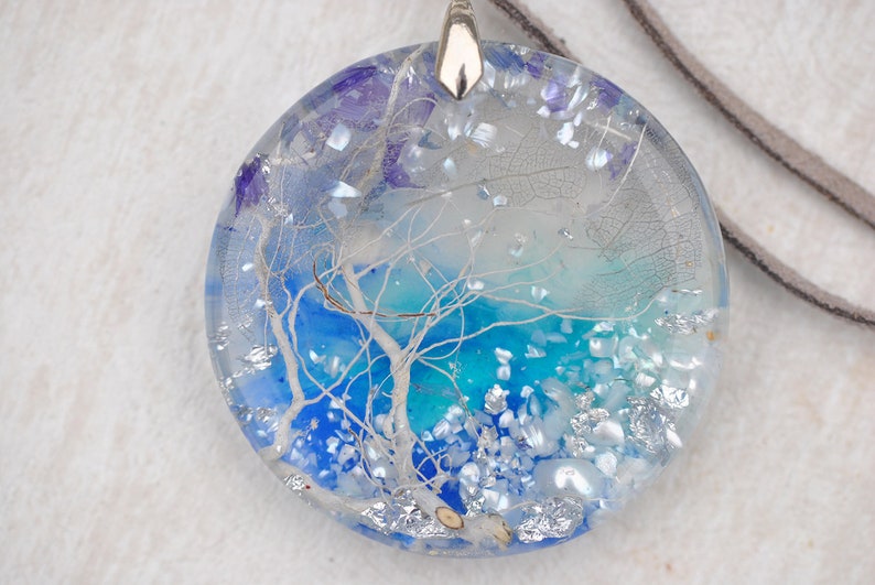 May include: A round, clear resin pendant with a blue and white tree design. The pendant is filled with silver glitter and white and blue iridescent flakes. The pendant is attached to a brown leather cord.