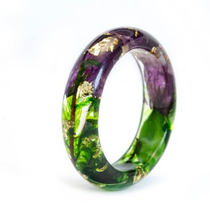 K&ouml;nnte beinhalten: Ein klarer Harzring mit eingebetteten lila Blumen, gr&uuml;nen Bl&auml;ttern und Goldflocken. Der Ring ist im Bandstil gehalten und hat eine glatte, polierte Oberfl&auml;che. Die floralen Elemente sind im gesamten Ring sichtbar und schaffen ein einzigartiges Accessoire.