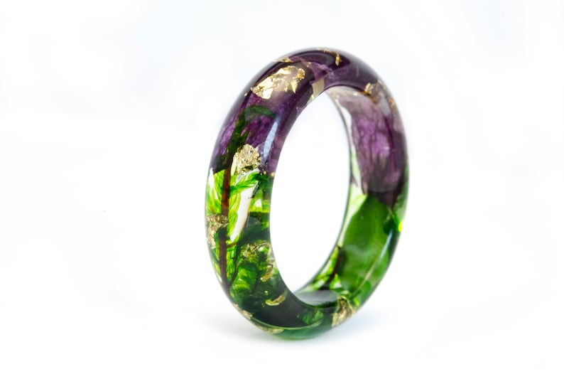 K&ouml;nnte beinhalten: Ein klarer Harzring mit eingebetteten lila Blumen, gr&uuml;nen Bl&auml;ttern und Goldflocken. Der Ring ist im Bandstil gehalten und hat eine glatte, polierte Oberfl&auml;che. Die floralen Elemente sind im gesamten Ring sichtbar und schaffen ein einzigartiges Accessoire.