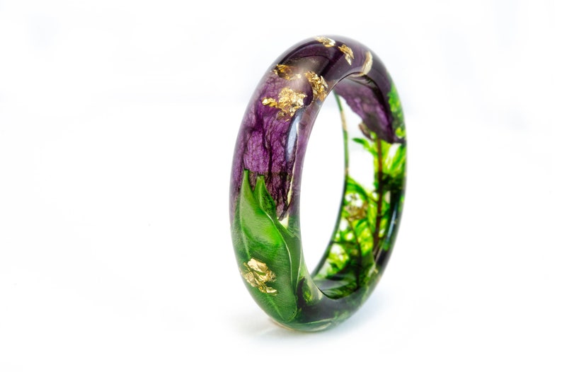 K&ouml;nnte beinhalten: Ein klarer Armreif aus Kunstharz mit lila Bl&uuml;ten, gr&uuml;nen Bl&auml;ttern und Goldblatt-Akzenten. Das botanische Design ist in dem transparenten Harz eingeschlossen und schafft ein einzigartiges Schmuckst&uuml;ck. Das Armband hat eine kreisf&ouml;rmige Form.