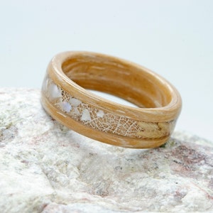 Può includere: Un anello in legno con una finitura marrone chiaro e una fascia di resina bianca con piccoli pezzi di conchiglia iridescente incorporati.