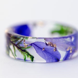 May include: A clear resin ring with embedded blue and green floral elements and gold flakes. The wide band ring showcases a botanical design, perfect for nature lovers. The ring's diameter is approximately 0.75 inches.