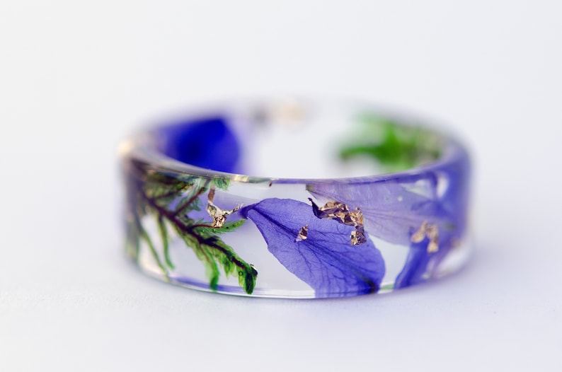 May include: A clear resin ring with embedded blue and green floral elements and gold flakes. The wide band ring showcases a botanical design, perfect for nature lovers. The ring's diameter is approximately 0.75 inches.
