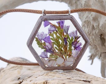 May include: A hexagonal pendant necklace with a brown wooden frame and a clear resin insert. The resin contains pressed purple flowers and white stones.