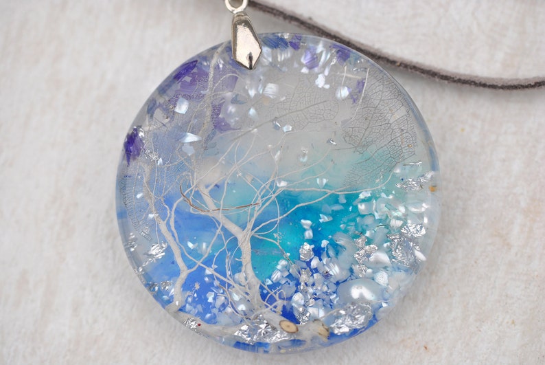 May include: A round, clear resin pendant necklace with a silver bail. The pendant contains a blue and white abstract design with silver glitter and white and blue iridescent flakes.