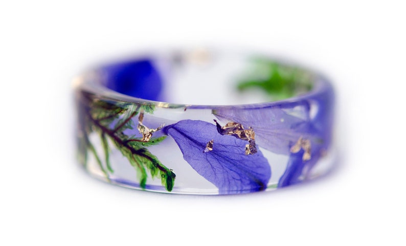 May include: A clear resin ring with embedded blue flower petals, green leaves, and gold flakes. The ring has a wide band and a smooth, polished finish. The floral elements are visible throughout the ring's transparent design.
