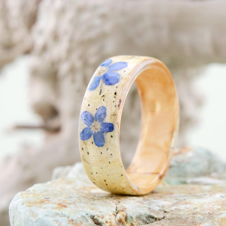 May include: A band ring featuring a smooth, light-colored, speckled surface embedded with two small, five-petaled blue flowers. The ring has a rounded profile and a polished finish, showcasing a natural, light-toned material throughout the band.