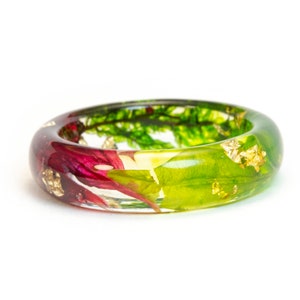 May include: A clear resin ring with embedded red, green, and yellow floral elements and gold leaf flakes. The ring has a rounded shape and is set against a white background.