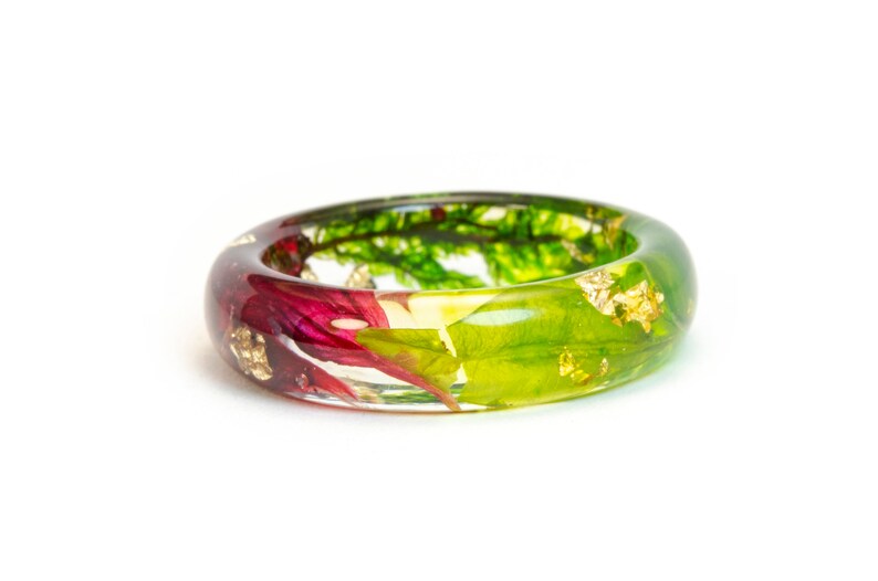 May include: A clear resin ring with embedded red, green, and yellow floral elements and gold leaf flakes. The ring has a rounded shape and is set against a white background.