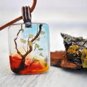 May include: A square pendant necklace with a brown leather cord. The pendant is made of clear resin with a small tree and leaves encased inside. The background of the pendant is a gradient of blue, orange, and red.