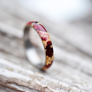 Puede incluir: Un fino anillo de banda de oro con una incrustaci&oacute;n roja y dorada. El anillo est&aacute; sobre una superficie de madera marr&oacute;n claro.