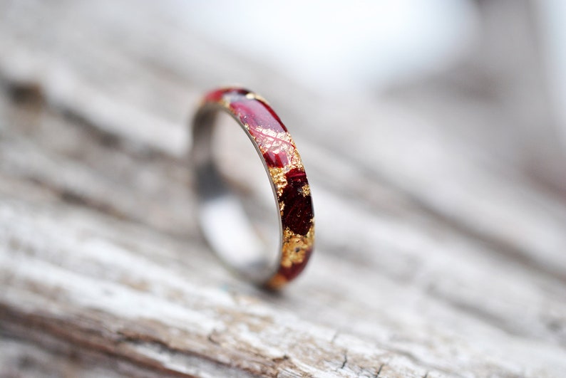 Puede incluir: Un fino anillo de banda de oro con una incrustaci&oacute;n roja y dorada. El anillo est&aacute; sobre una superficie de madera marr&oacute;n claro.