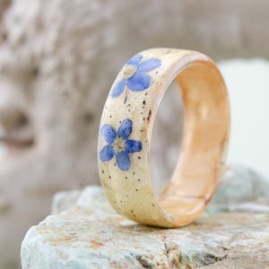 May include: A ring featuring a light beige band with a smooth, polished finish, decorated with two small, five-petaled blue flowers embedded into the surface. The interior of the band shows a natural, warm-toned wood grain texture and finish.