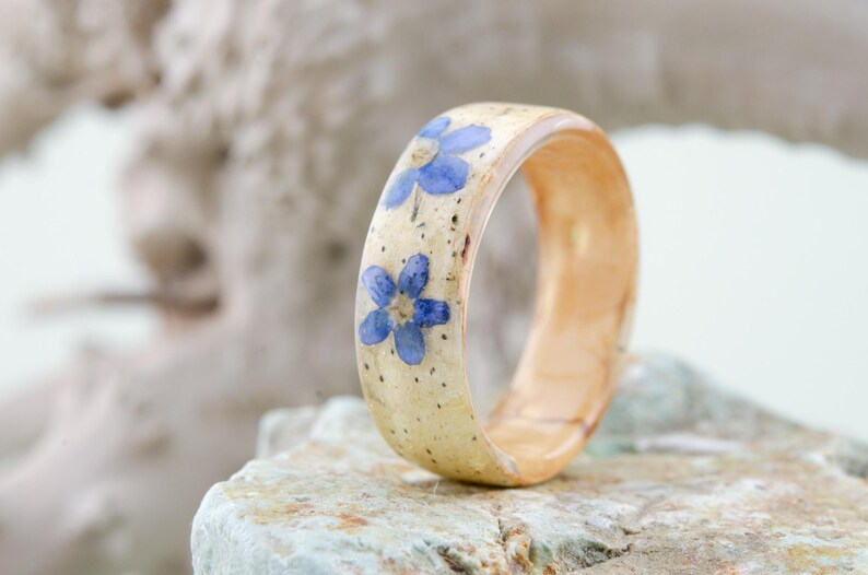 May include: A ring featuring a light beige band with a smooth, polished finish, decorated with two small, five-petaled blue flowers embedded into the surface. The interior of the band shows a natural, warm-toned wood grain texture and finish.