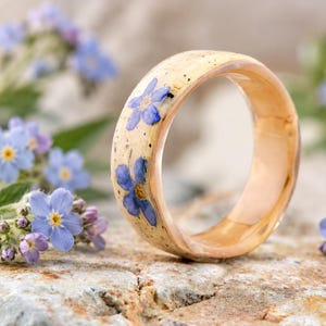 May include: A ring with a smooth, light-colored band featuring embedded blue forget-me-not flowers. The exterior surface of the ring has a speckled appearance, while the interior shows a polished, warm-toned wood grain finish.