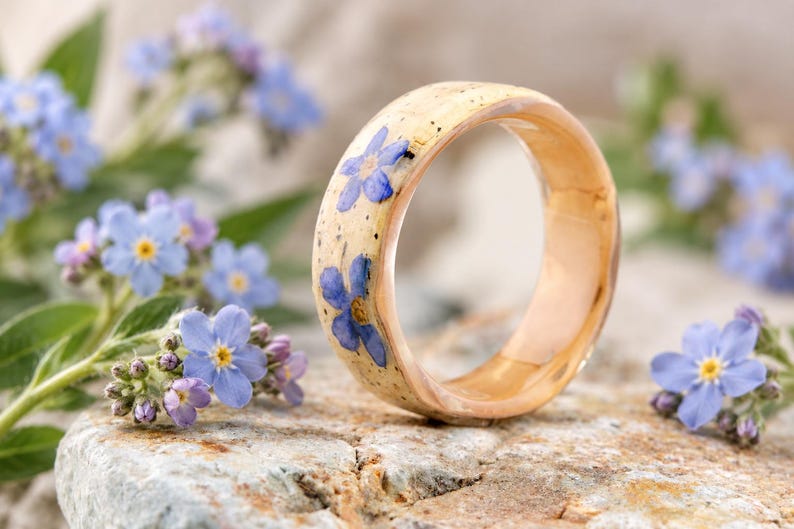 May include: A ring with a smooth, light-colored band featuring embedded blue forget-me-not flowers. The exterior surface of the ring has a speckled appearance, while the interior shows a polished, warm-toned wood grain finish.