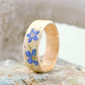 May include: A ring featuring a light beige band with a smooth, polished finish. The exterior surface is embedded with two small, vibrant blue five-petaled flowers and subtle dark speckles throughout the light-colored material of the band.