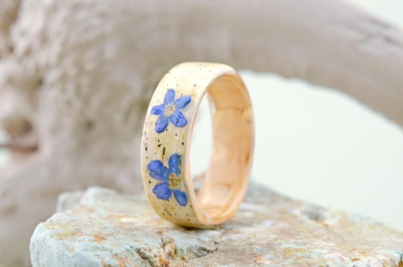 May include: A ring featuring a light beige band with a smooth, polished finish. The exterior surface is embedded with two small, vibrant blue five-petaled flowers and subtle dark speckles throughout the light-colored material of the band.