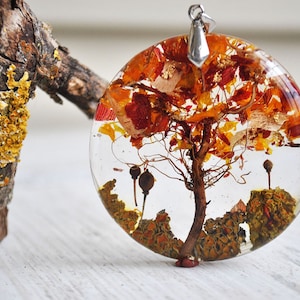 May include: A round, clear resin pendant with a small tree and dried flowers inside. The pendant is a light brown color with hints of orange and red. The pendant is hanging from a silver chain.