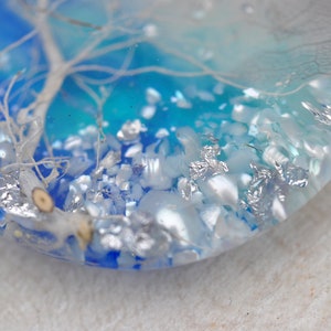 May include: A close-up of a round, clear resin pendant with a white tree branch and silver glitter embedded inside. The background is a gradient of blue and turquoise.