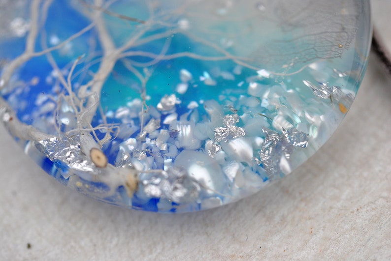 May include: A close-up of a round, clear resin pendant with a white tree branch and silver glitter embedded inside. The background is a gradient of blue and turquoise.