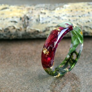 May include: A clear resin ring with embedded red and green floral elements and gold leaf flakes. The ring has a rounded shape and is set against a neutral background. The ring is a unique piece of jewelry.