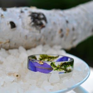 May include: A clear resin ring with embedded purple flower petals, green sprigs, and gold flakes. The ring is set on a bed of white crystals, with a blurred birch branch in the background. A unique, nature-inspired jewelry piece.