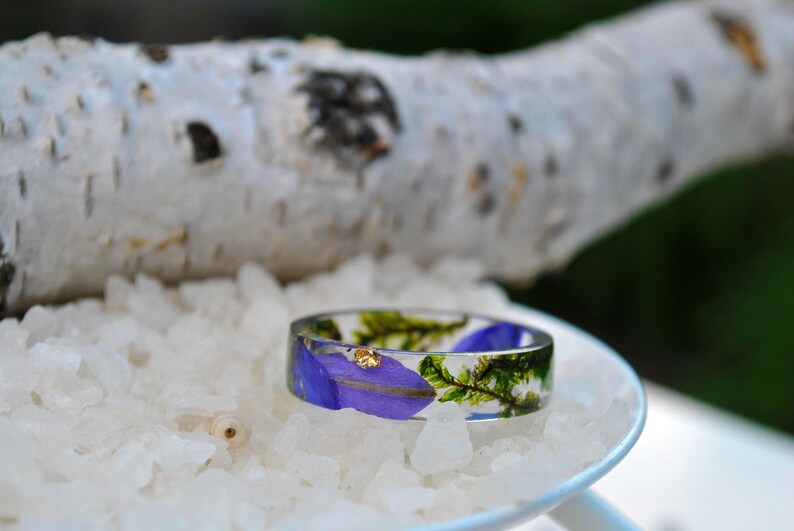May include: A clear resin ring with embedded purple flower petals, green sprigs, and gold flakes. The ring is set on a bed of white crystals, with a blurred birch branch in the background. A unique, nature-inspired jewelry piece.