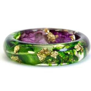 K&ouml;nnte beinhalten: Ein klarer Harzring mit eingebetteten gr&uuml;nen Bl&auml;ttern, lila Bl&uuml;ten und Goldflocken. Der Ring hat eine abgerundete Form und ist vor einem wei&szlig;en Hintergrund platziert. Ein einzigartiges, von der Natur inspiriertes Schmuckst&uuml;ck.