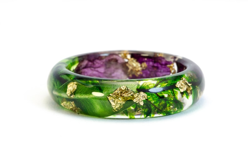 K&ouml;nnte beinhalten: Ein klarer Harzring mit eingebetteten gr&uuml;nen Bl&auml;ttern, lila Bl&uuml;ten und Goldflocken. Der Ring hat eine abgerundete Form und ist vor einem wei&szlig;en Hintergrund platziert. Ein einzigartiges, von der Natur inspiriertes Schmuckst&uuml;ck.