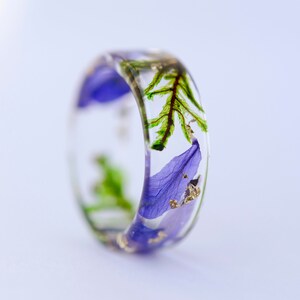 May include: A clear resin ring with embedded purple flower petals, green leaves, and gold flakes. The ring has a wide band and a smooth, polished finish. The floral design is visible from all angles.