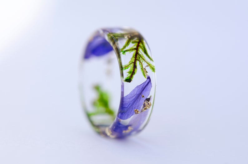 May include: A clear resin ring with embedded purple flower petals, green leaves, and gold flakes. The ring has a wide band and a smooth, polished finish. The floral design is visible from all angles.