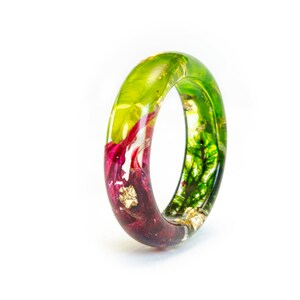 May include: A clear resin ring with embedded botanical elements. The ring features a gradient of colors, transitioning from deep burgundy to vibrant green. Gold leaf flakes add sparkle.