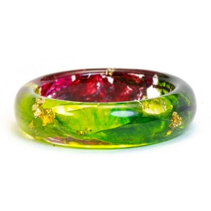 May include: A clear resin bangle bracelet with embedded red and green floral elements and gold leaf accents. The bracelet has a rounded shape and is set against a white background. The colors are vibrant and the design is unique.