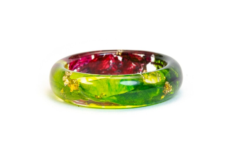 May include: A clear resin bangle bracelet with embedded red and green floral elements and gold leaf accents. The bracelet has a rounded shape and is set against a white background. The colors are vibrant and the design is unique.