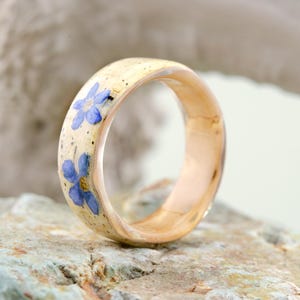 May include: A ring with a smooth, light-colored band featuring embedded blue forget-me-not flowers and small dark speckles. The interior of the band is a polished, warm-toned metallic finish, showcasing a rounded profile and a clear, glossy surface.