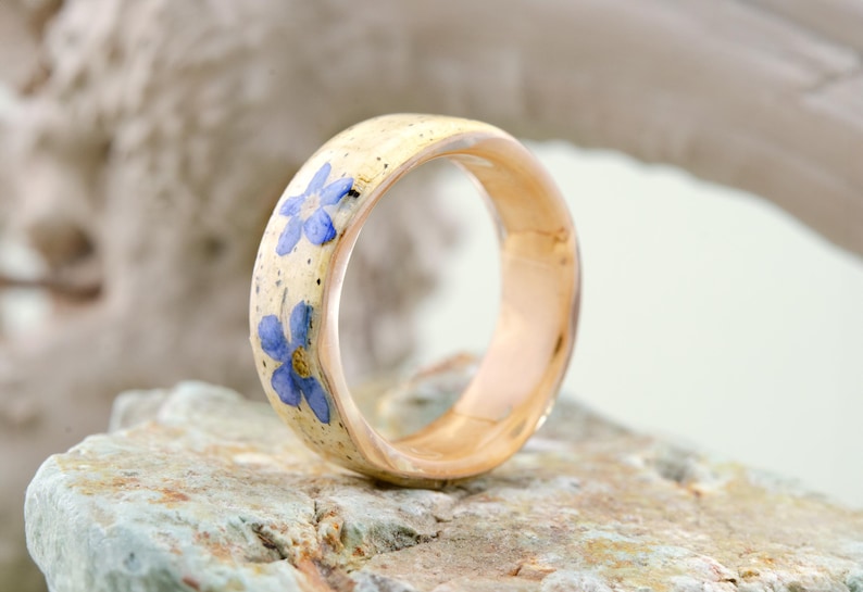 May include: A ring with a smooth, light-colored band featuring embedded blue forget-me-not flowers and small dark speckles. The interior of the band is a polished, warm-toned metallic finish, showcasing a rounded profile and a clear, glossy surface.