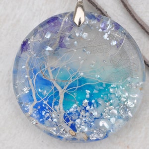 May include: A round, clear resin pendant necklace with a blue and white abstract design. The pendant features a white tree branch with a blue and turquoise background. The necklace has a silver chain and a brown leather cord.