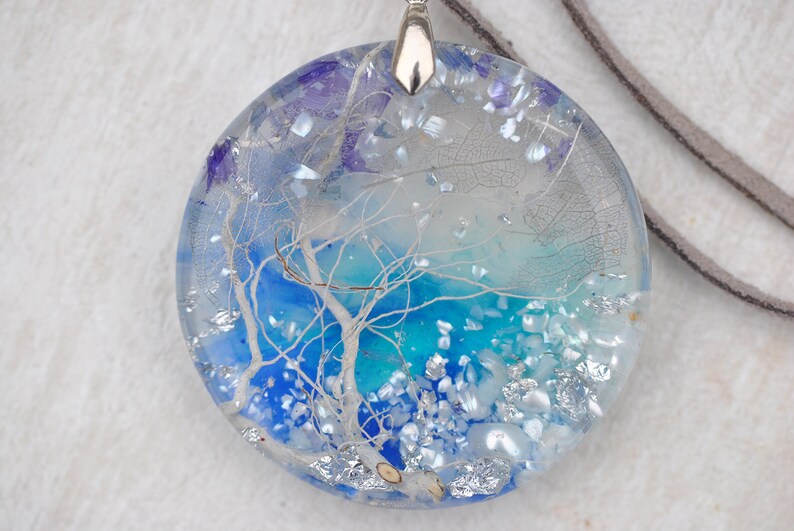 May include: A round, clear resin pendant necklace with a blue and white abstract design. The pendant features a white tree branch with a blue and turquoise background. The necklace has a silver chain and a brown leather cord.