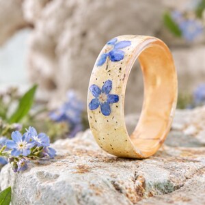 May include: A band ring featuring a smooth, light beige, speckled surface embedded with small, vibrant blue five-petaled flowers. The ring has a polished, rounded exterior and a warm, light-toned interior finish, showcasing a natural aesthetic.